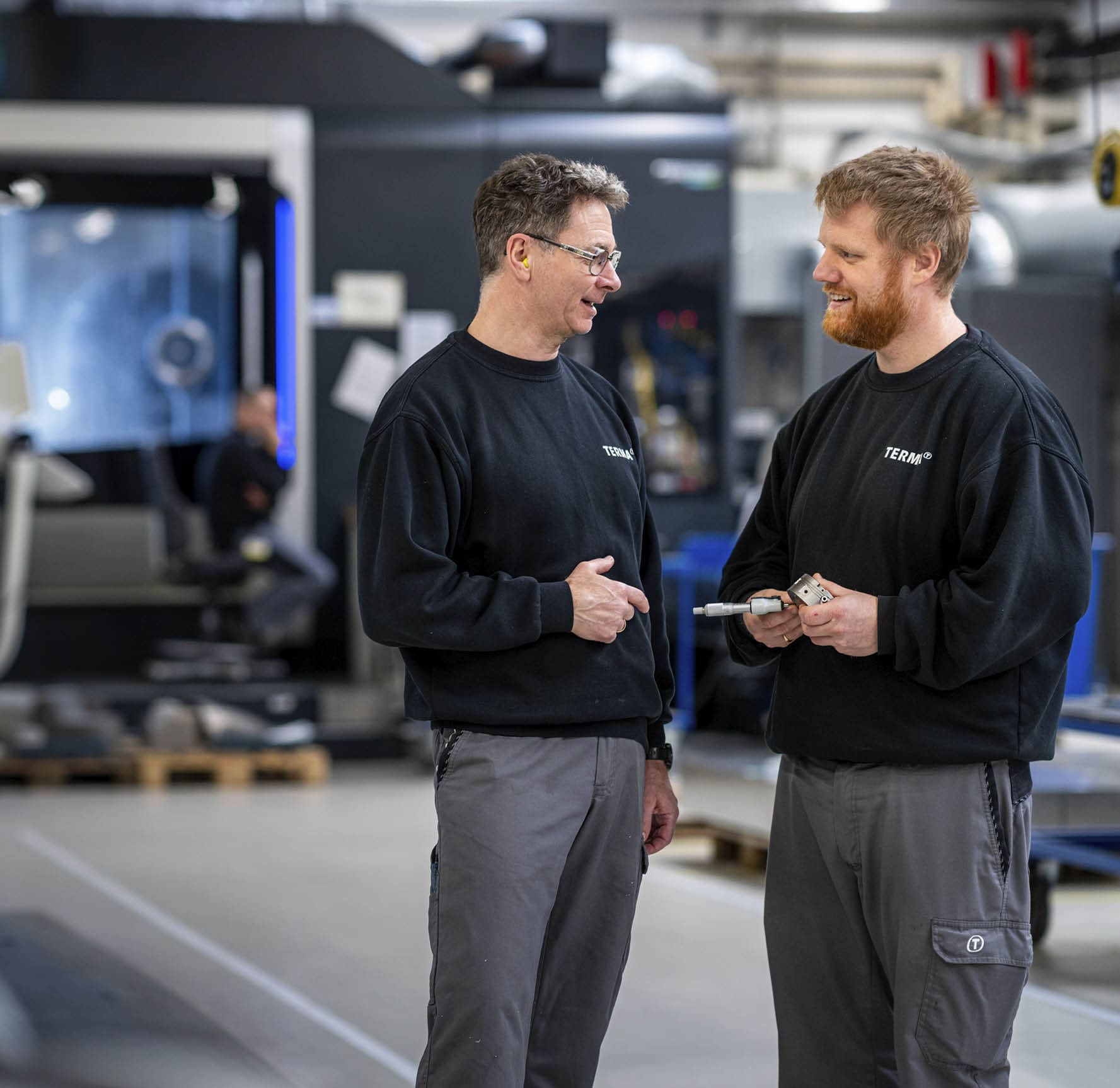 Two adults wearing black sweatshirts and gray pants are standing and talking in an industrial workshop with machinery and equipment in the background. Description generated by AI