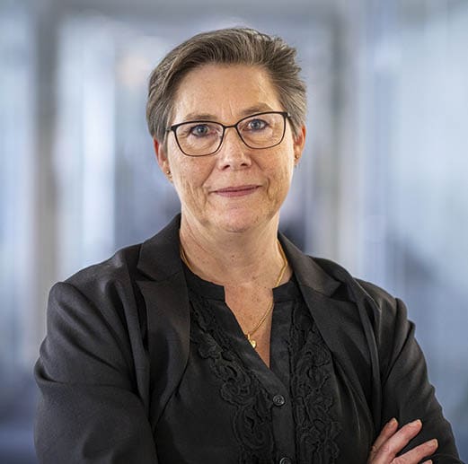 A woman wearing a black jacket and glasses is standing in an office with her arms crossed. AI generated content
