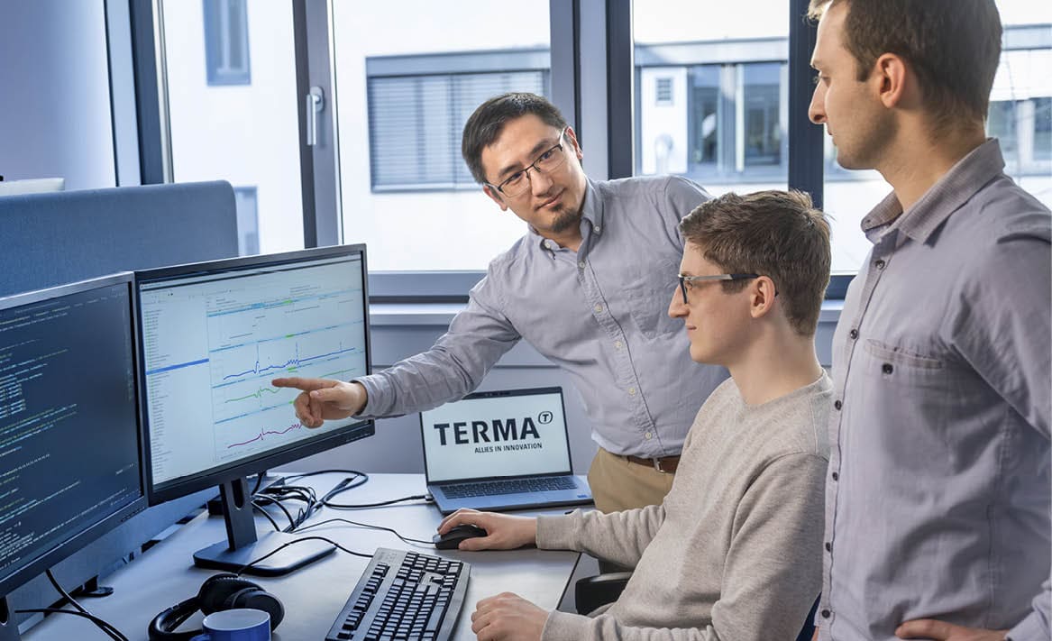Three men are standing around a desk with a laptop and a monitor. One man is pointing at the laptop. AI generated content