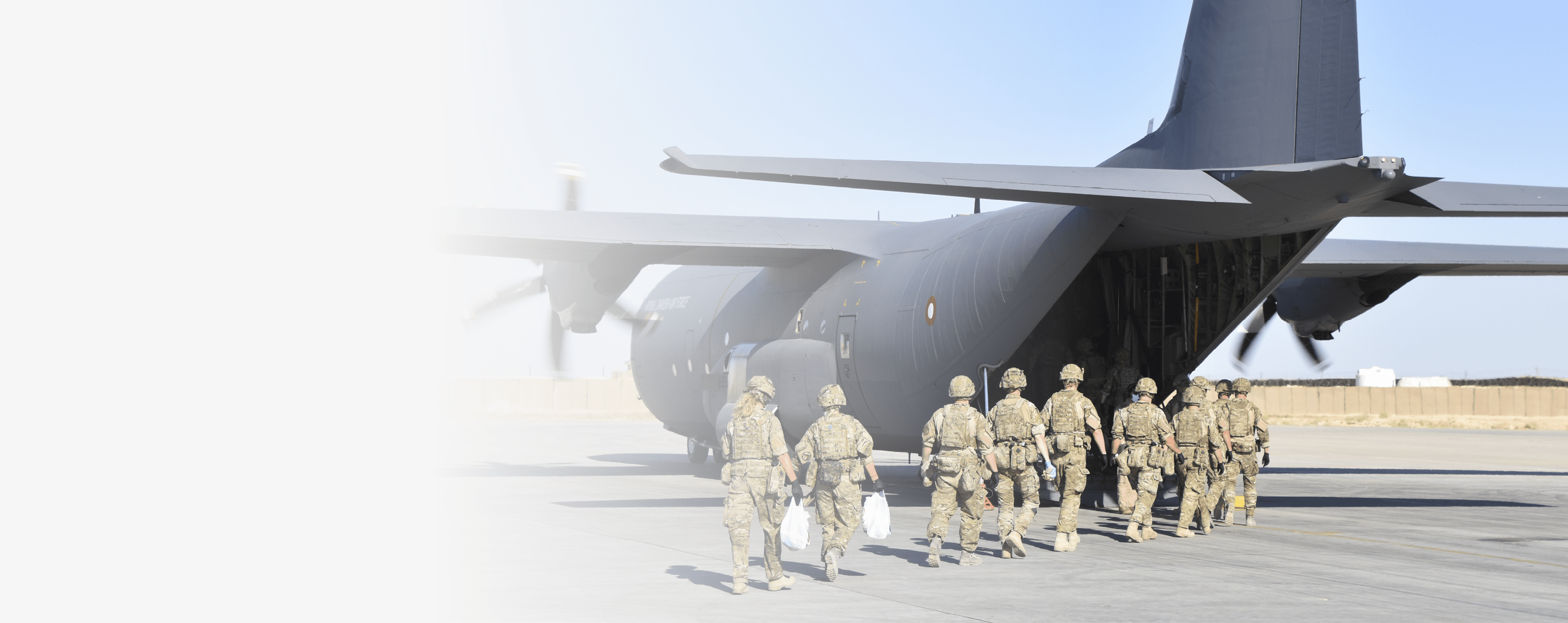 A group of people in military uniforms walk towards the open rear cargo door of a large gray military transport aircraft on a sunny airfield. Description generated by AI