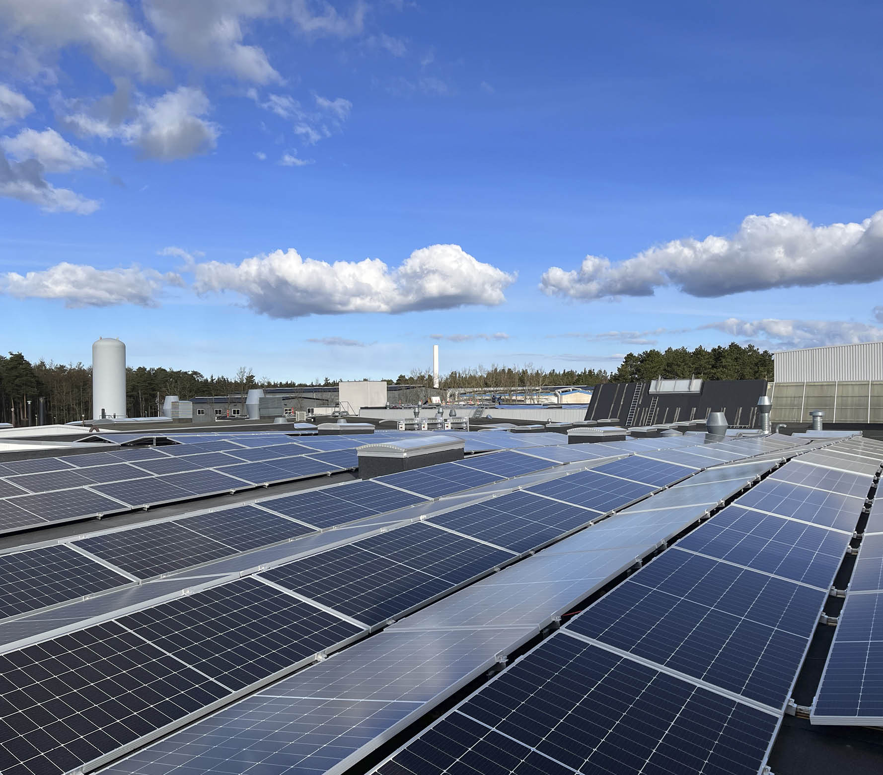 Rows of solar panels are installed on a flat rooftop under a partly cloudy blue sky with trees and industrial buildings in the background. Description generated by AI