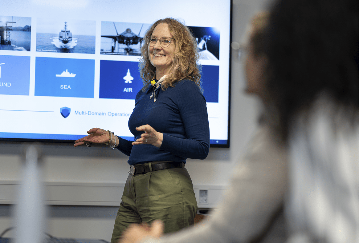 A person with curly hair wearing a blue long-sleeve shirt and green pants is giving a presentation in front of a screen displaying categories labeled ground, sea, and air while another person listens in the foreground. Description generated by AI