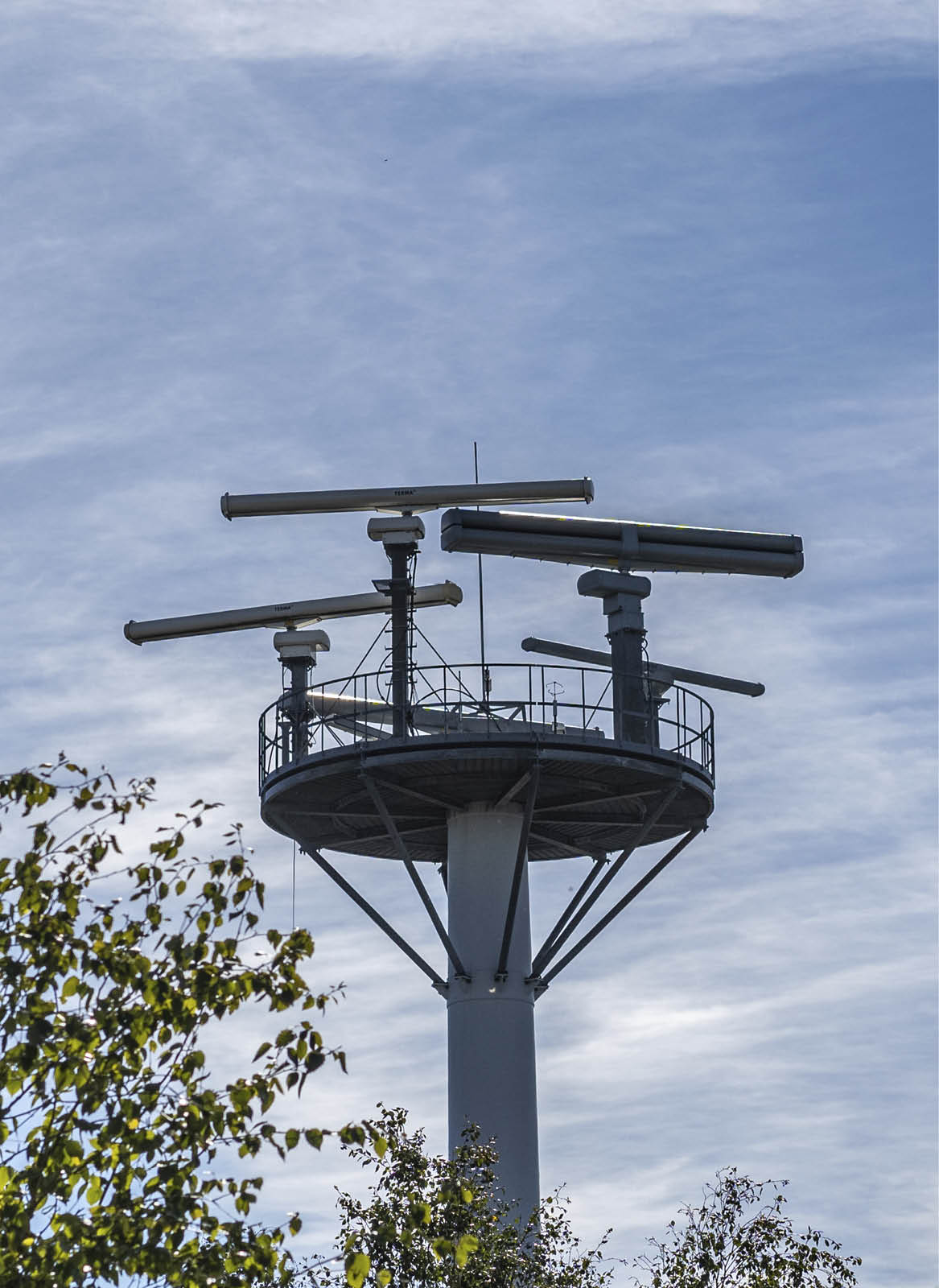 A tall metal structure with antennas stands behind a leafy tree and a brick building under a partly cloudy blue sky. Description generated by AI